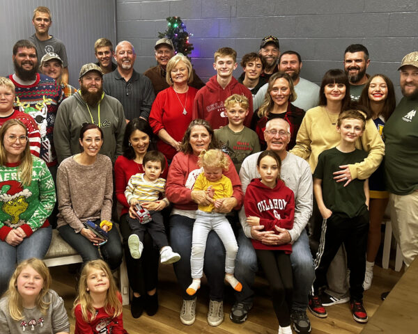 Large family gathering with diverse group of adults and children, festive attire, and a small Christmas tree in the background, celebrating together.